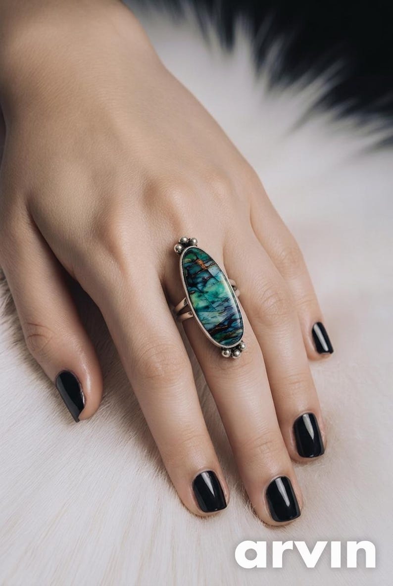May include: A sterling silver ring featuring an elongated oval-shaped stone with swirling patterns of deep green, teal, and dark brown. The metal setting includes small decorative silver spheres at the top and bottom of the stone, with a split band.