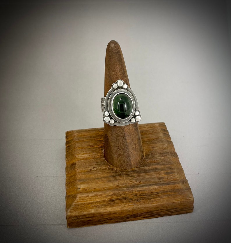 May include: A silver ring with a green gemstone set in a triangular bezel. The ring is decorated with small silver beads around the bezel.
