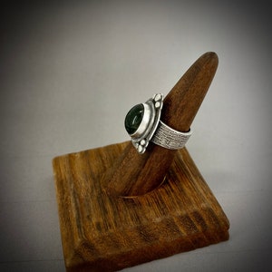 May include: A silver ring with a dark green gemstone set in a textured band. The ring is displayed on a wooden ring holder.
