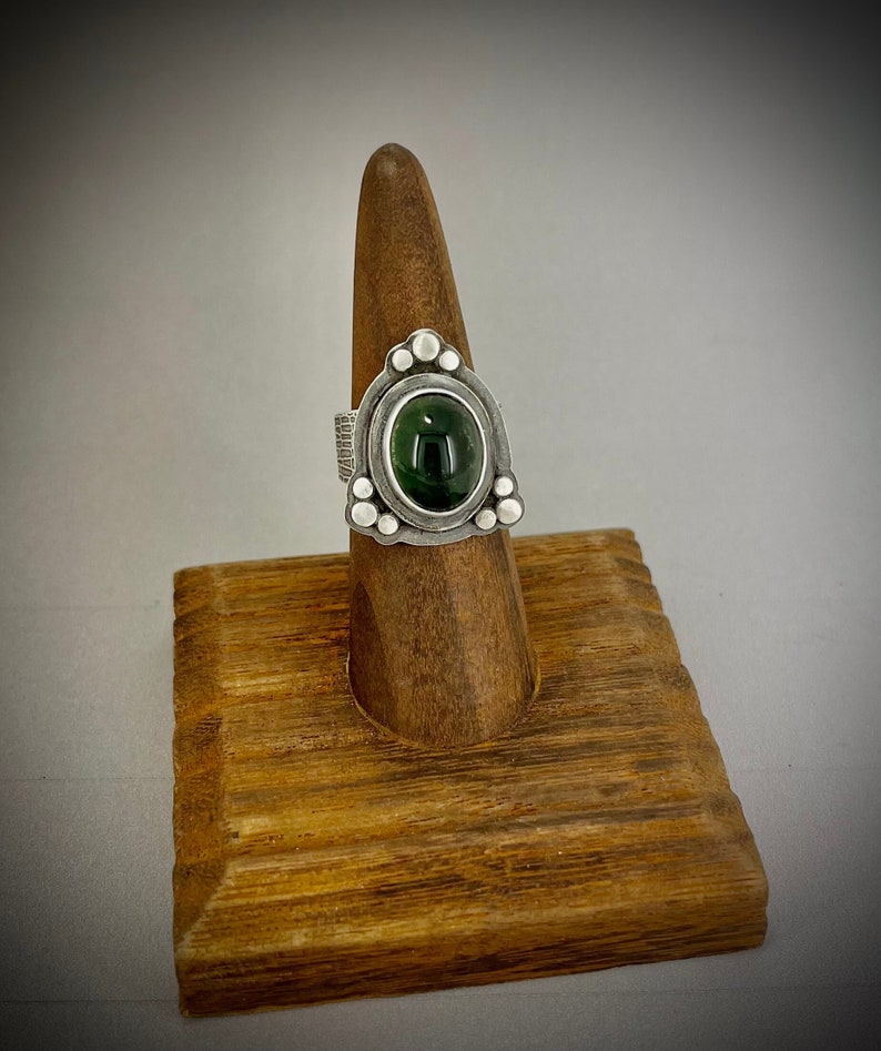 May include: A silver ring with a green gemstone set in a triangular bezel. The ring is on a wooden ring display stand.