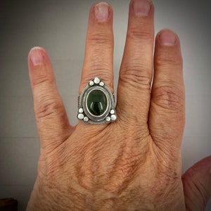 May include: A silver ring with a green oval gemstone set in a triangular bezel. The ring is decorated with small silver beads around the bezel.