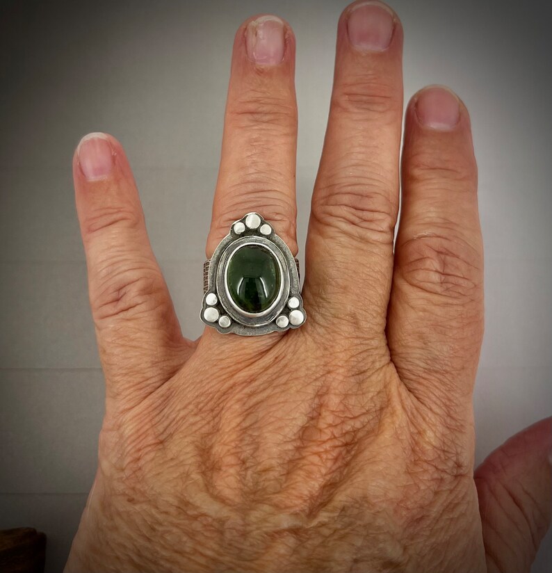 May include: A silver ring with a green oval gemstone set in a triangular bezel. The ring is decorated with small silver beads around the bezel.