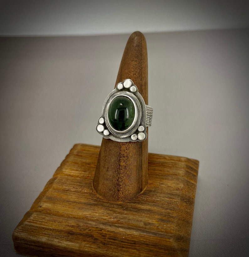 May include: A silver ring with a green gemstone set in a triangular bezel. The ring is decorated with small silver beads around the bezel.