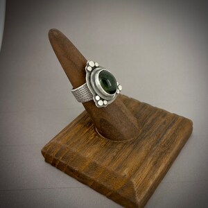 May include: A silver ring with a green gemstone set in a textured band. The ring is on a wooden ring display stand.