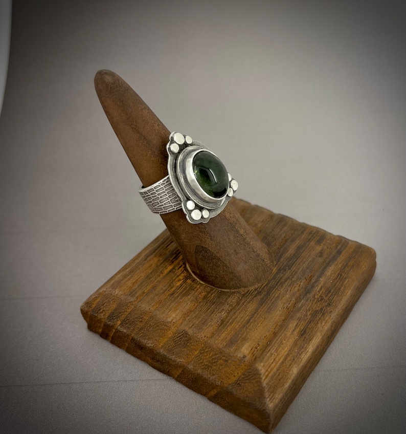 May include: A silver ring with a green gemstone set in a textured band. The ring is on a wooden ring display stand.