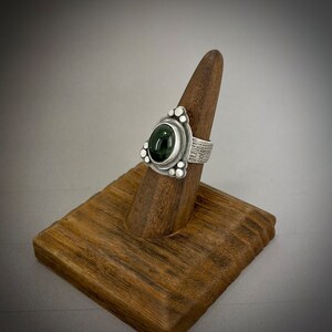 May include: A silver ring with a dark green gemstone set in a triangular bezel. The band is textured and has small silver beads around the bezel.