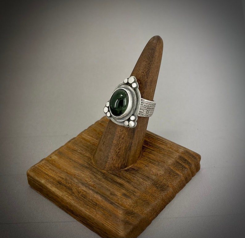 May include: A silver ring with a dark green gemstone set in a triangular bezel. The band is textured and has small silver beads around the bezel.