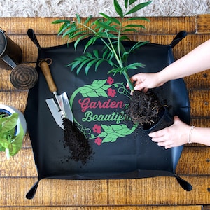 May include: A black gardening mat with the words "Garden Beautiful" printed in green and red. A person is repotting a plant with brown roots onto the mat. The mat has a raised edge to prevent soil from spilling.