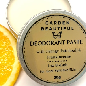 May include: A silver tin with a gold label that reads "GARDEN BEAUTIFUL DEODORANT PASTE with Orange, Patchouli & Frankincense Low Bi-Carb for more Sensitive Skin 30g". A butterfly is on the label. A slice of orange is in the background.