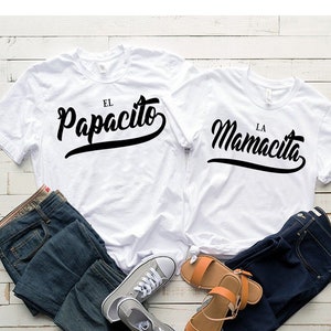 Puede incluir: Dos camisetas blancas con texto negro. La camiseta de la izquierda dice "El Papacito" y la camiseta de la derecha dice "La Mamacita". Las camisetas est&aacute;n dispuestas sobre una superficie de madera blanca con un par de pantalones vaqueros azules y un par de zapatillas de deporte a cada lado.