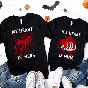 May include: Two black t-shirts with a Halloween theme. The left shirt says "My heart is hers" with a bloody heart. The right shirt says "His heart is mine" with a skeleton hand holding a bloody heart.