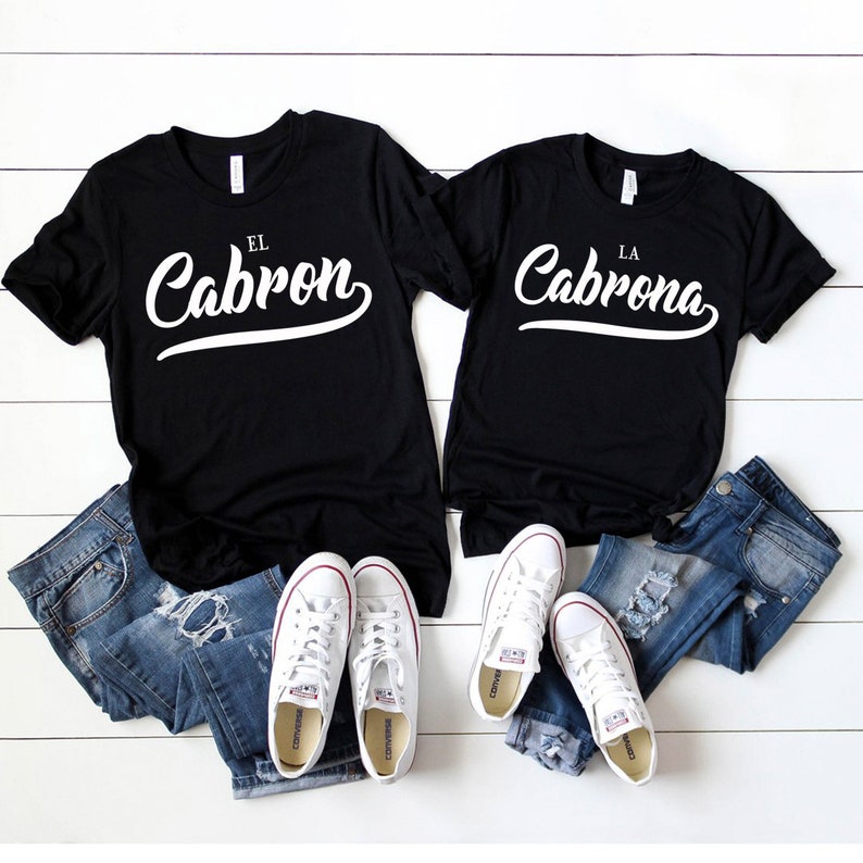 Puede incluir: Dos camisetas negras con texto blanco. La camiseta de la izquierda dice "El Cabron" y la camiseta de la derecha dice "La Cabrona". Las camisetas est&aacute;n dispuestas sobre una superficie blanca con pantalones vaqueros azules y zapatillas blancas.