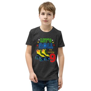 May include: Dark grey t-shirt with a colourful graphic that reads "Let's Roll, I'm turning 9." The graphic features a pair of yellow and blue roller skates. The shirt is worn by a person.