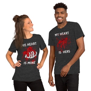 May include: Two dark grey t-shirts with a graphic print of a bloody heart. The text on the left shirt reads "His heart is mine". The text on the right shirt reads "My heart is hers".