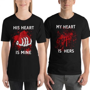 May include: Two black t-shirts with a graphic print of a bloody heart. The text on the first shirt reads "His heart is mine". The text on the second shirt reads "My heart is hers".