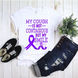 Puede incluir: Camiseta blanca con una cinta morada y texto que dice "My cough... is not... contagious but my smile is". El texto "CF Warrior" está escrito en la cinta.