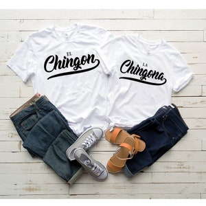 Puede incluir: Dos camisetas blancas con texto negro. La camiseta de la izquierda dice "El Chingon" y la camiseta de la derecha dice "La Chingona". Las camisetas est&aacute;n dispuestas sobre una superficie de madera blanca con un par de pantalones vaqueros azules y un par de zapatillas grises.