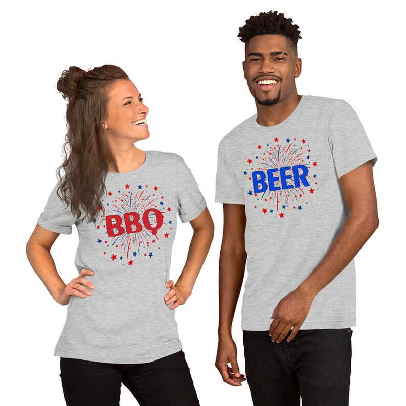May include: Two grey t-shirts with red, white, and blue firework designs. One shirt says "BBQ" and the other says "BEER".