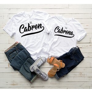 Puede incluir: Dos camisetas blancas con texto negro. La camiseta de la izquierda dice "El Cabron" y la camiseta de la derecha dice "La Cabrona". Las camisetas est&aacute;n dispuestas sobre una superficie de madera blanca con un par de pantalones vaqueros azules y un par de sandalias marrones.