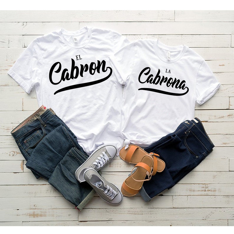 Puede incluir: Dos camisetas blancas con texto negro. La camiseta de la izquierda dice "El Cabron" y la camiseta de la derecha dice "La Cabrona". Las camisetas est&aacute;n dispuestas sobre una superficie de madera blanca con un par de pantalones vaqueros azules y un par de sandalias marrones.