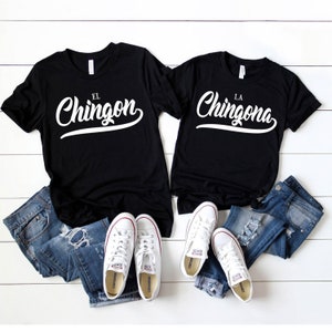 Puede incluir: Dos camisetas negras con texto blanco. La camiseta de la izquierda dice "El Chingon" y la camiseta de la derecha dice "La Chingona". Las camisetas est&aacute;n dispuestas sobre una superficie de madera blanca con jeans azules y zapatillas blancas.