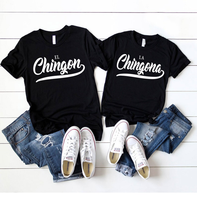 Puede incluir: Dos camisetas negras con texto blanco. La camiseta de la izquierda dice "El Chingon" y la camiseta de la derecha dice "La Chingona". Las camisetas est&aacute;n dispuestas sobre una superficie de madera blanca con jeans azules y zapatillas blancas.