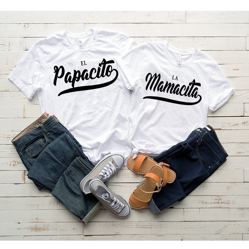 Puede incluir: Dos camisetas blancas con texto negro. La camiseta de la izquierda dice "El Papacito" y la camiseta de la derecha dice "La Mamacita". Las camisetas est&aacute;n dispuestas sobre una superficie de madera blanca con un par de pantalones vaqueros azules y un par de zapatillas de deporte a cada lado.