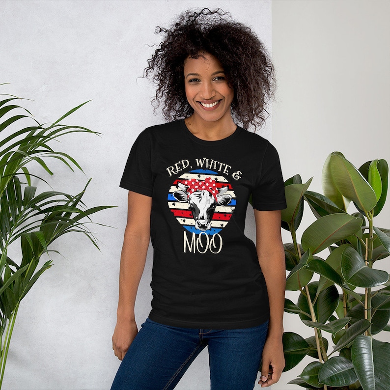 Może przedstawiać: Czarny T-shirt z grafiką czerwono-biało-niebieskiej krowy. Krowa nosi czerwoną, białą i niebieską chustkę. Na koszulce nadrukowany jest napis "Red, White & Moo".