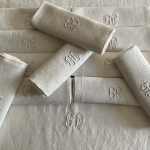 Puede incluir: Un conjunto de servilletas de lino blanco, cada una bordada con las letras "GE" en una escritura decorativa. Las servilletas están dobladas y dispuestas sobre una superficie blanca, mostrando su textura y diseño.