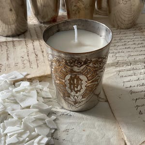 Peut inclure: Un bougeoir en argent avec un motif doré orné et une bougie blanche à l'intérieur. Le bougeoir est posé sur un morceau de papier avec du texte manuscrit.