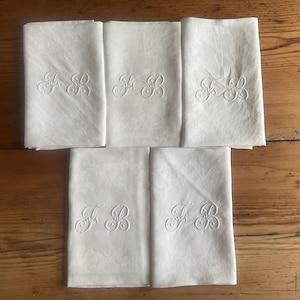 Puede incluir: Un conjunto de seis servilletas de lino blanco, cada una doblada y con un monograma "FB" bordado. Las servilletas están dispuestas sobre una superficie de madera, mostrando su aspecto impecable y su diseño clásico. Ideales para cenas formales u ocasiones especiales. Cada servilleta mide aproximadamente 40 cm x 40 cm.