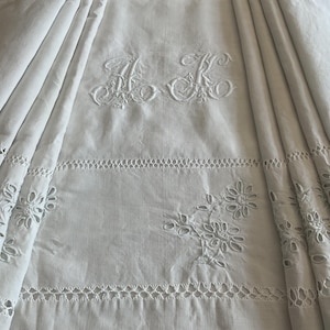 May include: White linen bedding with embroidered initials and floral cutwork. The bedding features decorative stitching and eyelet details. The initials "A K" are embroidered in a decorative script.