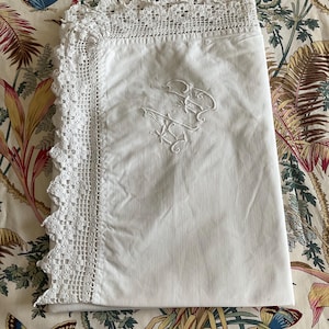 May include: A folded white linen pillowcase with a crocheted lace border. The pillowcase features embroidered initials in the center. The background is a floral patterned fabric with butterflies.