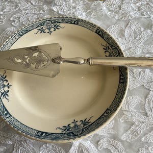 Puede incluir: Un servidor de pastel vintage descansa sobre un plato crema con un borde floral azul. El servidor es plateado con un diseño calado adornado en la hoja y un mango acanalado. El plato tiene aproximadamente 23 cm de diámetro.