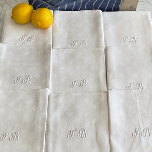 May include: A set of eight white fabric napkins arranged in a grid, each featuring a subtle tonal floral pattern and the embroidered monogram SB in cursive script. Two bright yellow lemons are placed on the top left napkin.