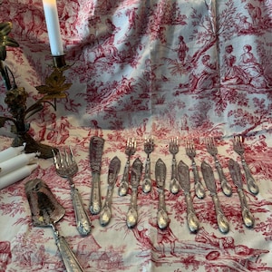 Puede incluir: Un conjunto de cubiertos plateados que incluye tenedores, cuchillos y un utensilio de servir, dispuestos sobre una tela blanca con un patrón de toile de Jouy rojo. Un candelabro de metal con una vela blanca encendida se sitúa a la izquierda.