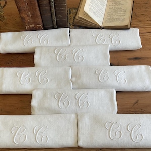 May include: Eight white linen napkins, each embroidered with the letters "CC". The napkins are rectangular and folded, arranged on a wooden surface. Antique books and decorative elements are in the background.