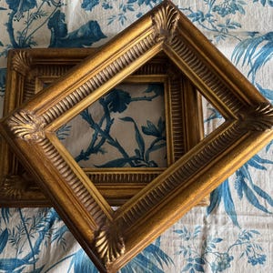 May include: Two rectangular picture frames with a metallic gold finish. Each frame features a raised, ribbed inner border and prominent shell-like decorative motifs at each of the four corners. The frames are stacked at an angle.
