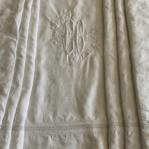 Peut inclure: Linge de lit en lin blanc avec un monogramme brodé au centre. Le linge de lit présente une bordure décorative et des plis verticaux. Le monogramme est entouré de broderies florales.