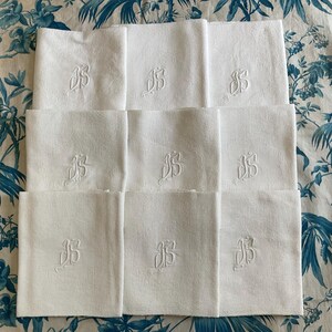May include: A set of nine white square napkins arranged in a three-by-three grid. Each napkin features a centered, tone-on-tone embroidered monogram of the letters JS in a cursive script, surrounded by a subtle, woven geometric border pattern.