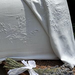 Può includere: Un tessuto bianco caratterizzato da un intricato ricamo floreale bianco con cuciture dettagliate. Il tessuto è drappeggiato, mostrando un delicato motivo botanico di fiori e steli. Un mazzetto di lavanda essiccata con un nastro bianco è posto sotto.