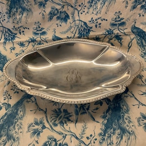 Può includere: Un piatto da portata ovale color argento con tre scomparti. Il piatto ha un bordo perlato e un monogramma al centro. Lo sfondo è un motivo floreale blu e bianco.