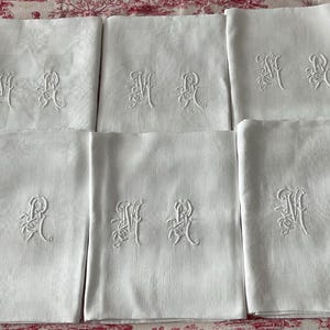 Può includere: Sei tovaglioli di lino bianco con iniziali ricamate. Ogni tovagliolo presenta un monogramma delicato e ornato in filo bianco coordinato. I tovaglioli sono piegati con cura e disposti su una superficie a fantasia, mostrando il loro design elegante.