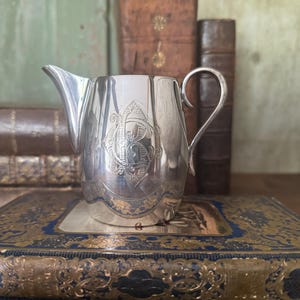 Peut inclure: Un pot à lait en argent poli doté d'une anse incurvée, d'un bec verseur pointu et d'un monogramme gravé complexe sur le devant. Le corps du pot présente un motif festonné décoratif à la base avec des gravures florales détaillées.