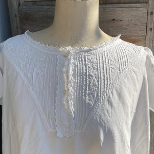 Puede incluir: Un camisón blanco vintage exhibido en un maniquí. El camisón presenta intrincados detalles de bordado y pliegues alrededor del escote y el corpiño. La prenda tiene mangas cortas y una parte delantera abotonada, mostrando un diseño delicado y femenino.