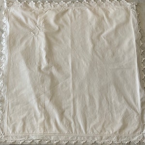 Puede incluir: Mantel de mesa de algodón blanco con un delicado borde de encaje y un pequeño diseño bordado en el centro.