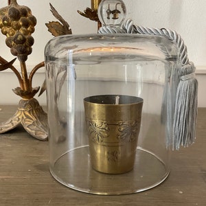 Può includere: Un portacandele in ottone con dettagli floreali è sotto una campana di vetro trasparente. Il portacandele ha uno stoppino acceso. Un nappa e una corda decorativi sono attaccati alla campana. Un oggetto decorativo color oro è sullo sfondo.