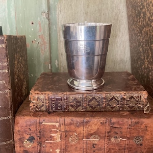 Peut inclure: Une coupe argentée avec un motif décoratif, posée sur une pile de livres anciens reliés en cuir. La coupe a un bord évasé et une base détaillée. Les livres sont de tailles variées et ont des accents dorés.