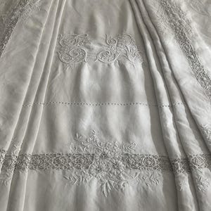 Può includere: Tovaglia di lino bianca con intricati ricami floreali e a volute. La tovaglia presenta bordi dettagliati e motivi centrali, creando un'estetica delicata ed elegante. Il tessuto è drappeggiato, mettendo in risalto la trama e l'artigianato.