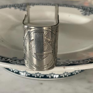 May include: A silver napkin ring with an engraved design, resting on a white plate with a blue floral border. The napkin ring has a rectangular panel and a curved top.